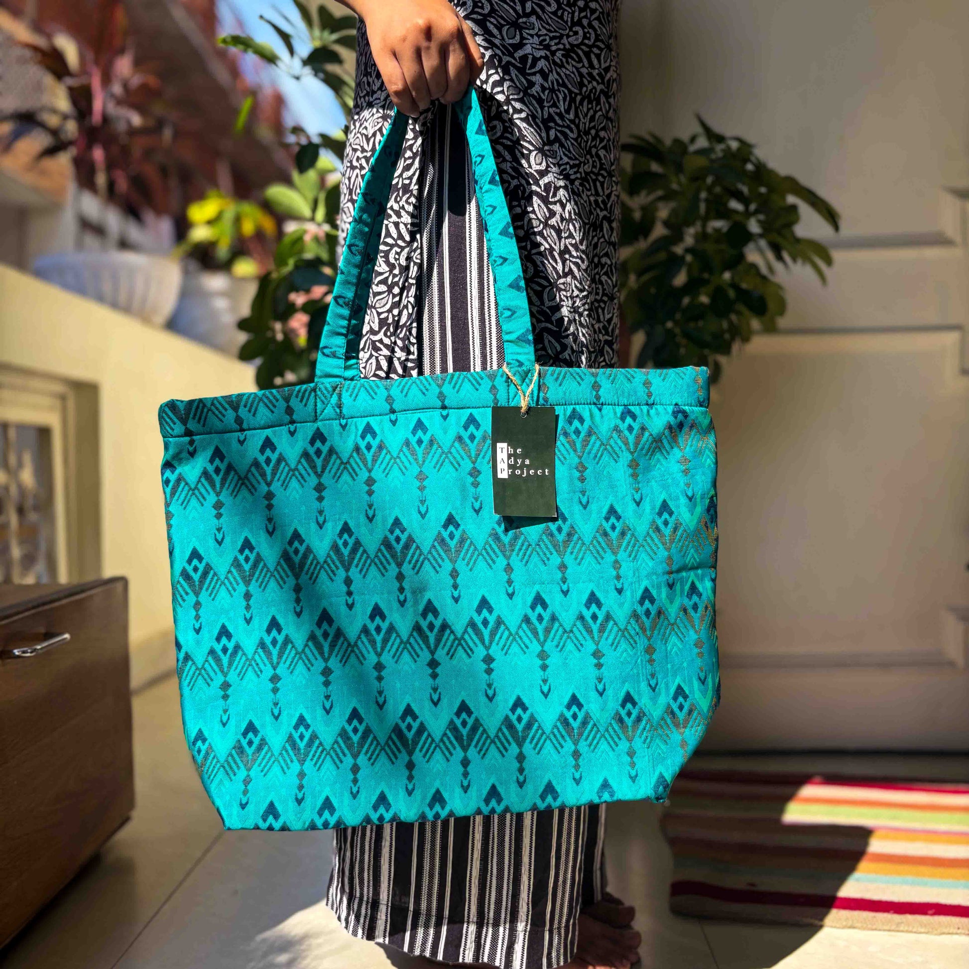 Person holding a turquoise ikat-patterned tote bag with a visible brand tag, standing indoors.