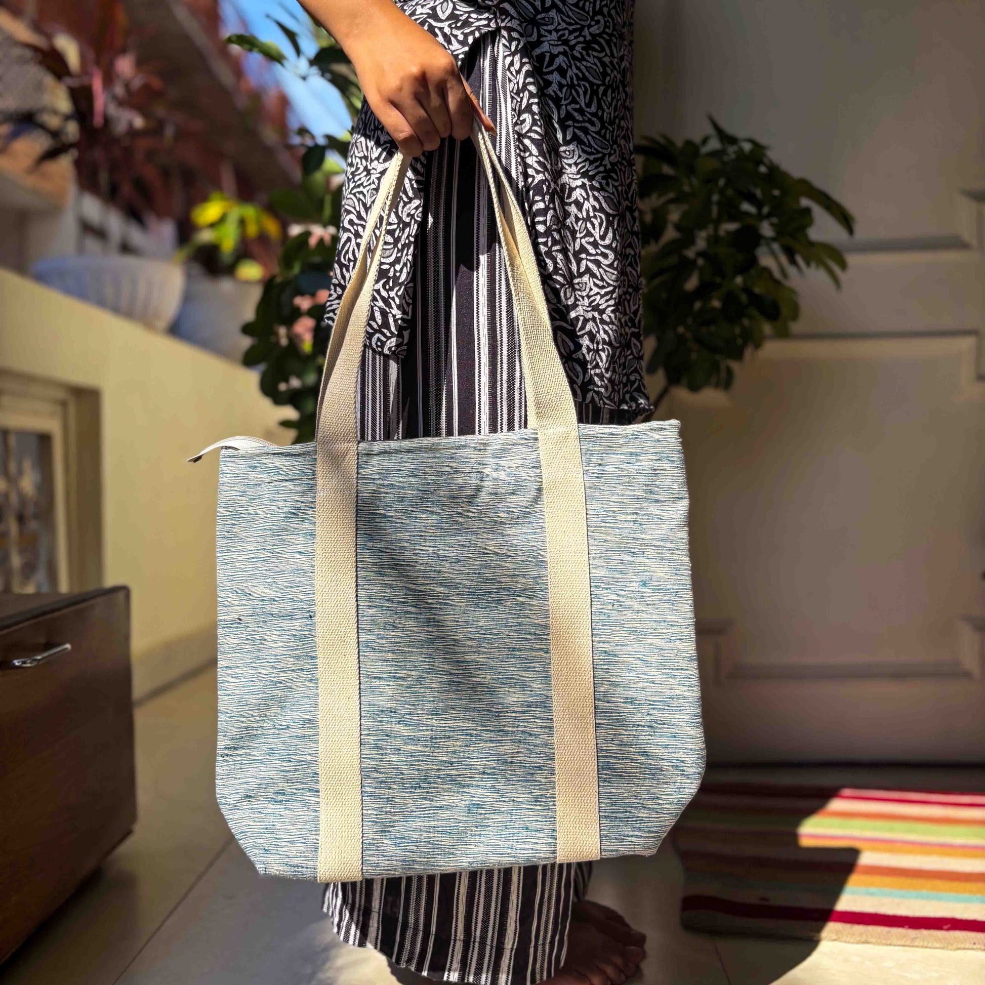 Person holding a blue tote bag in a home setting with plants and furniture.