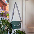 Green handbag with a lace strap hanging on a white door next to green plants.