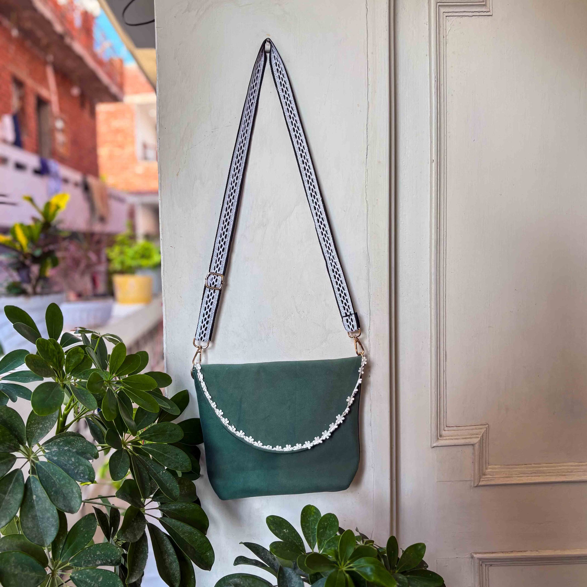 Green handbag with a lace strap hanging on a white door next to green plants.