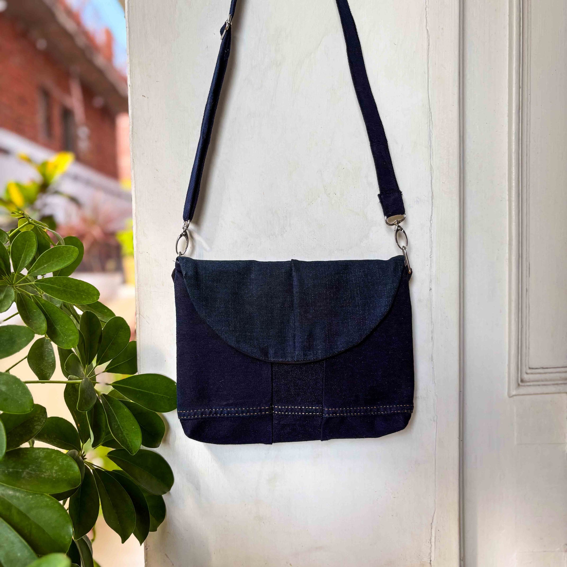 Navy blue crossbody bag hanging on a white door with a plant in the foreground.