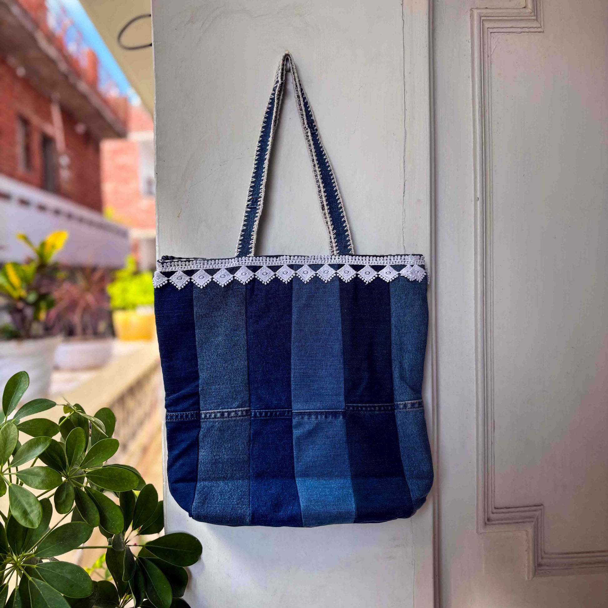 Blue denim tote bag with white lace trim hanging on a white door.