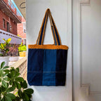 Blue and orange tote bag hanging on a white door with plants and a building in the background.