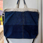 Blue denim tote bag with crochet top and zipper, hanging on a light-colored wall.