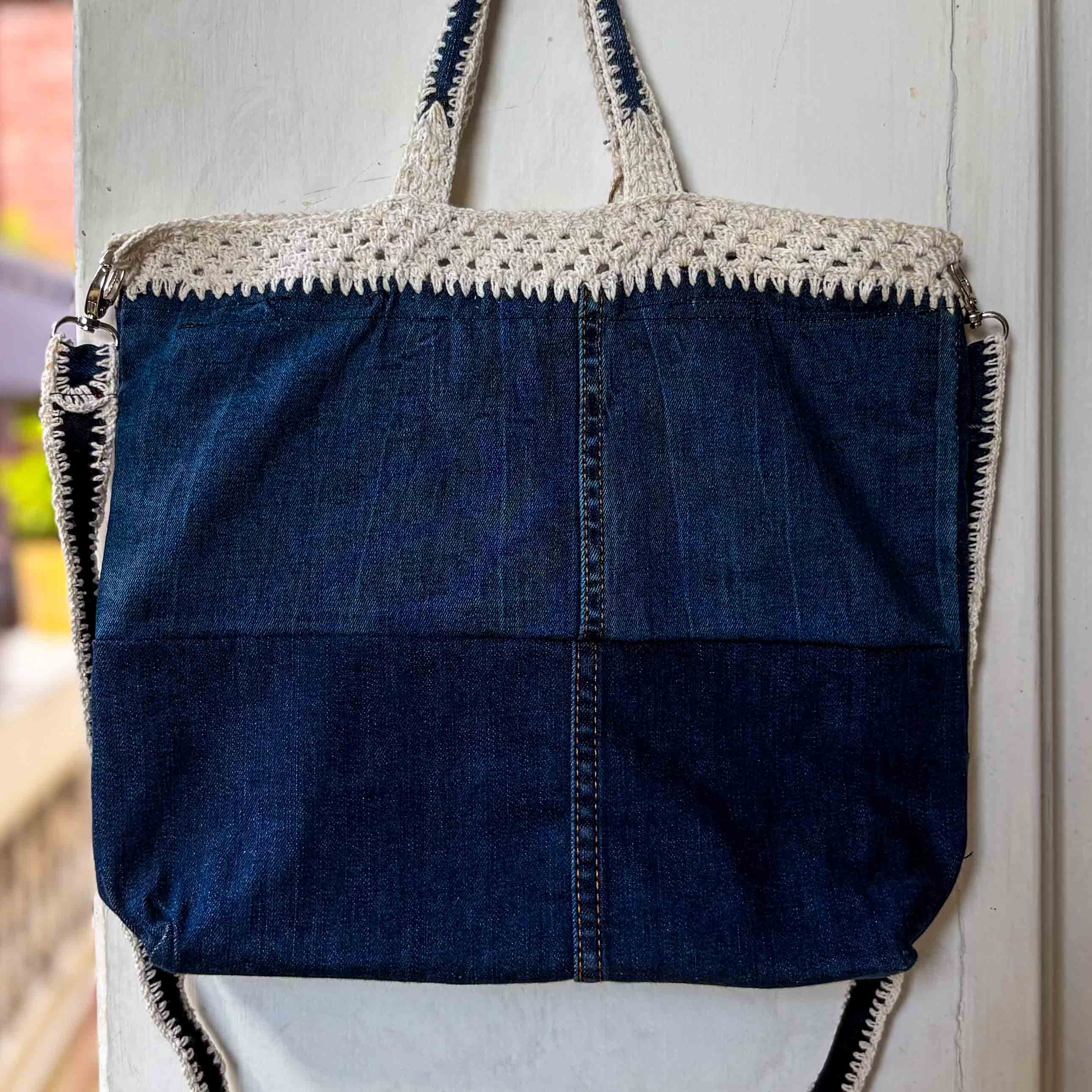 Blue denim tote bag with crochet top and zipper, hanging on a light-colored wall.