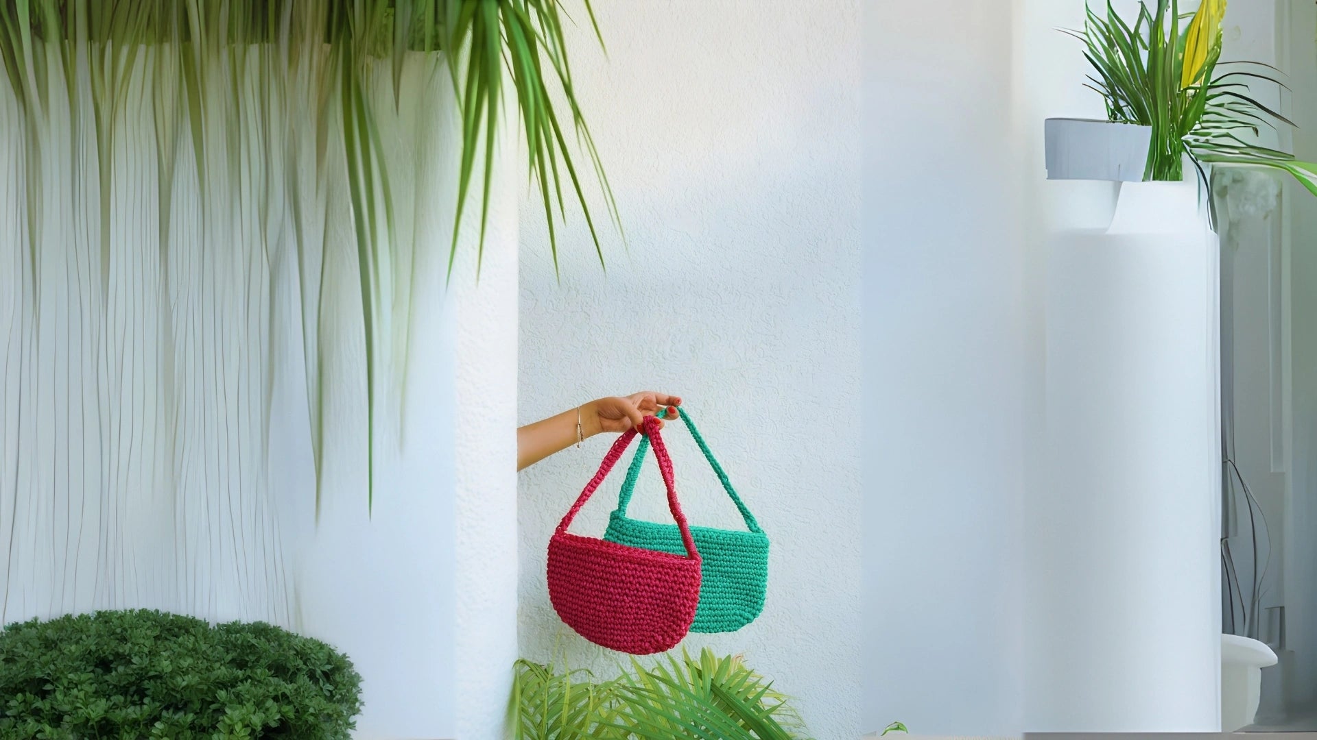 Two woven handbags, one red and one green, held by a person against a white wall with plants.